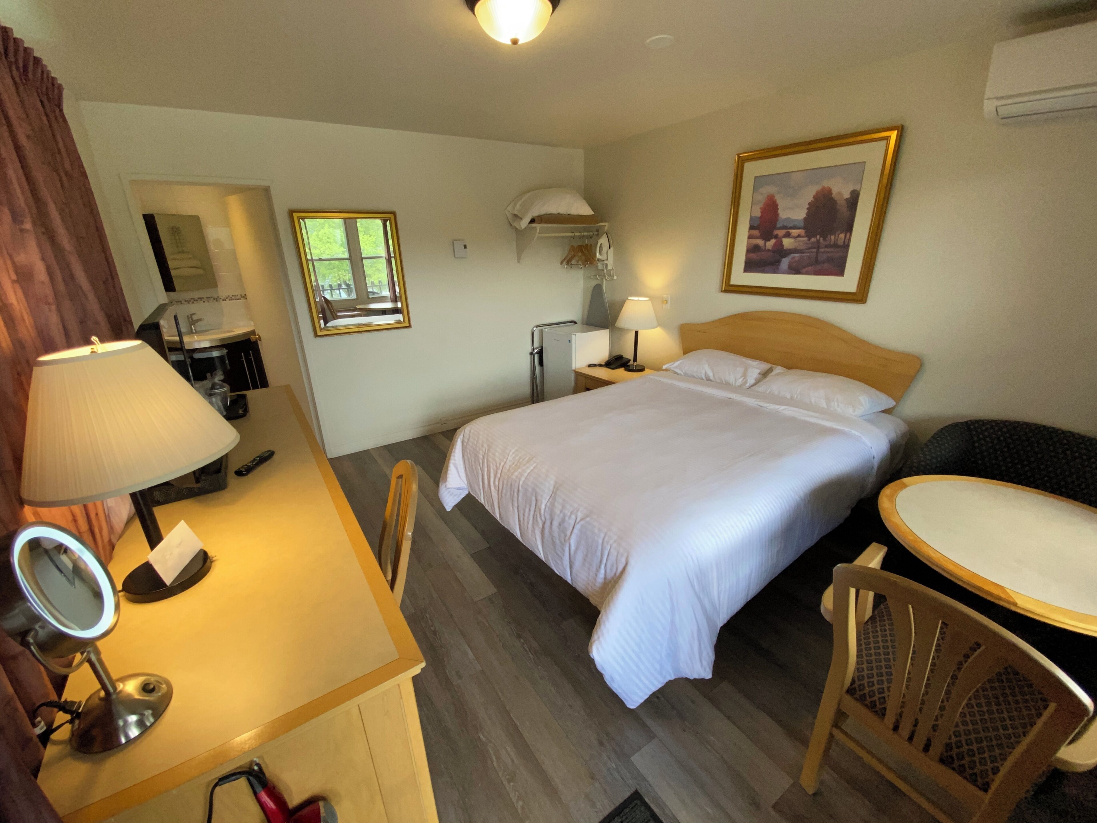 A hotel room with a bed, bedside table, chair, desk, lamp, mirror, and a painting on the wall.