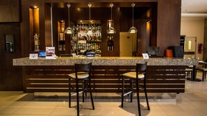 A bar area with a granite countertop, multiple hanging pendant lights, and a selection of liquor bottles. There are two bar stools and a small table with a menu. - Carnaval Hotel Casino (Encarnacion)