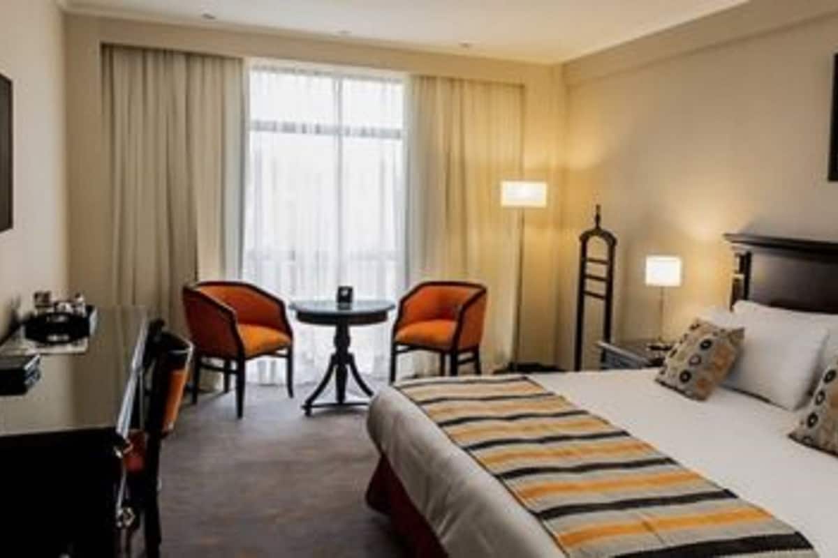 A hotel room with a bed, two chairs, a small table, a desk, a TV, and a painting on the wall.