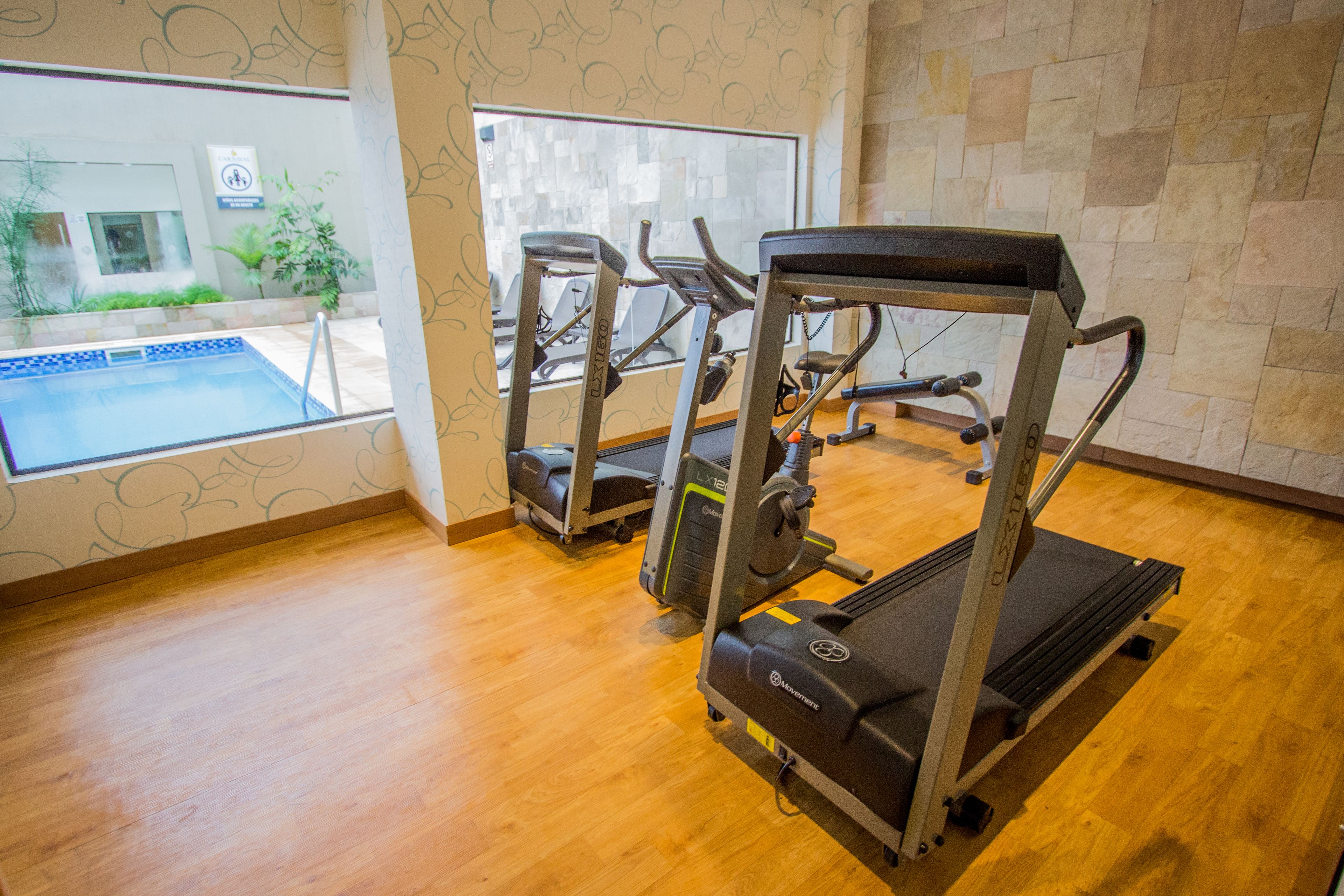 Une salle de sport équipée de tapis de course et d’une piscine visible à travers les fenêtres.