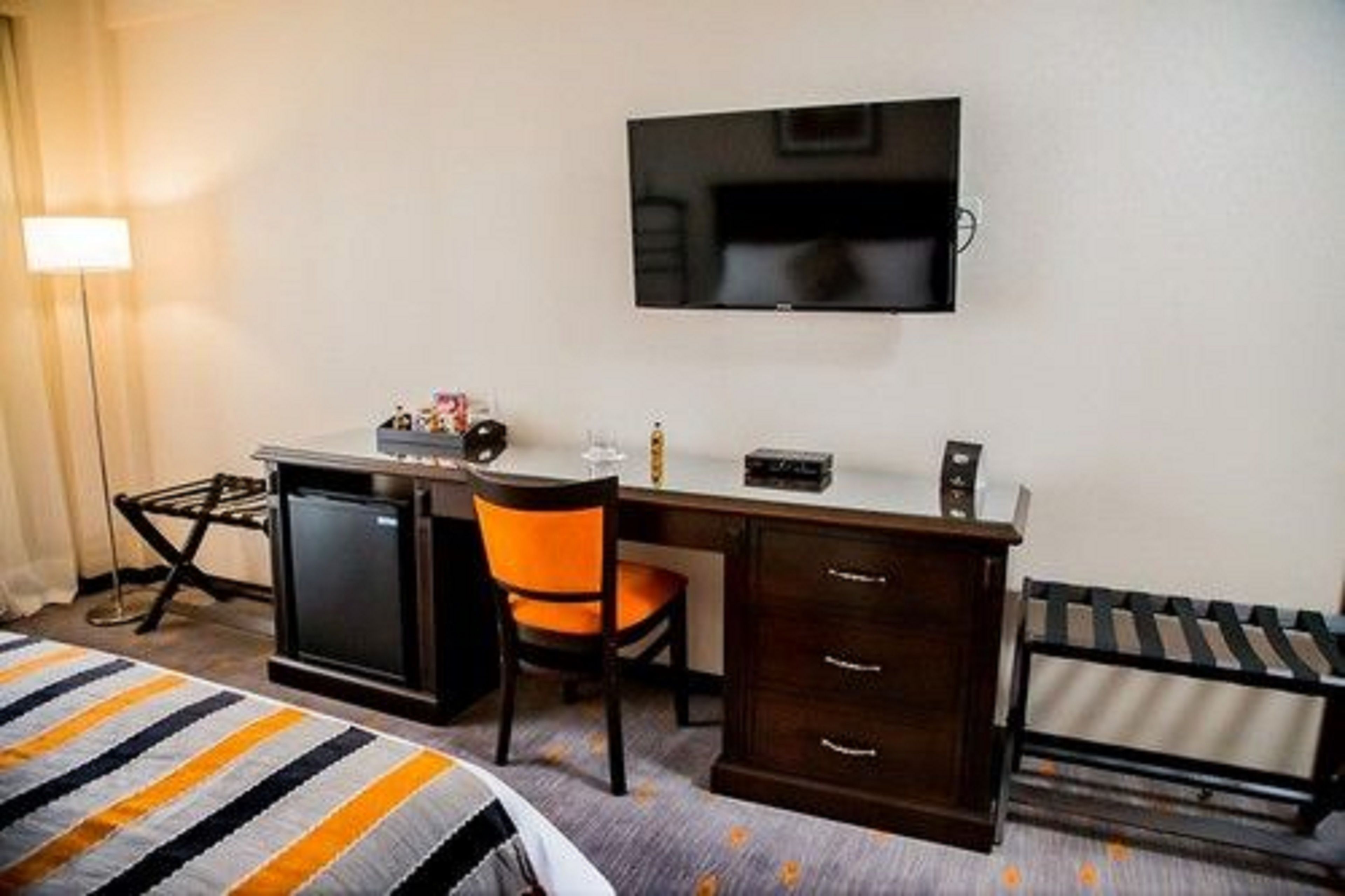 A hotel room with a bed, a desk with a chair, a television mounted on the wall, and a small refrigerator.