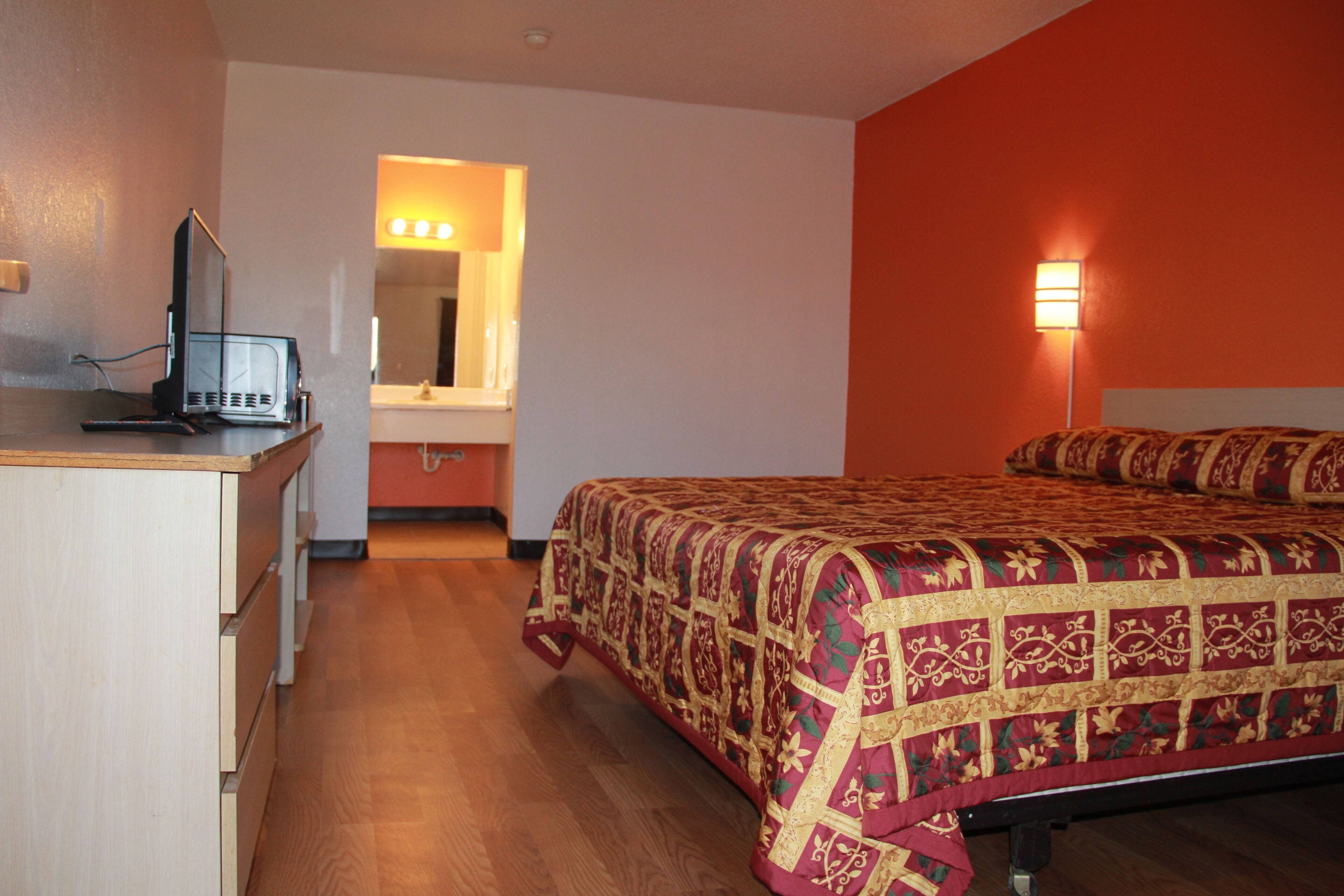A hotel room with a bed, a desk with a computer, a mirror, and a wall-mounted light.