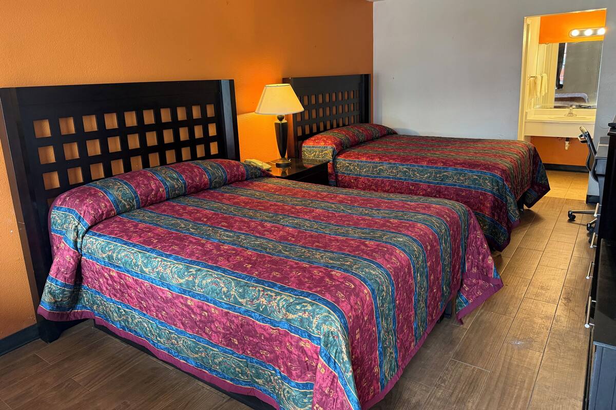 A hotel room with two beds, a wooden headboard, a nightstand with a lamp, and a bathroom with a mirror and sink.