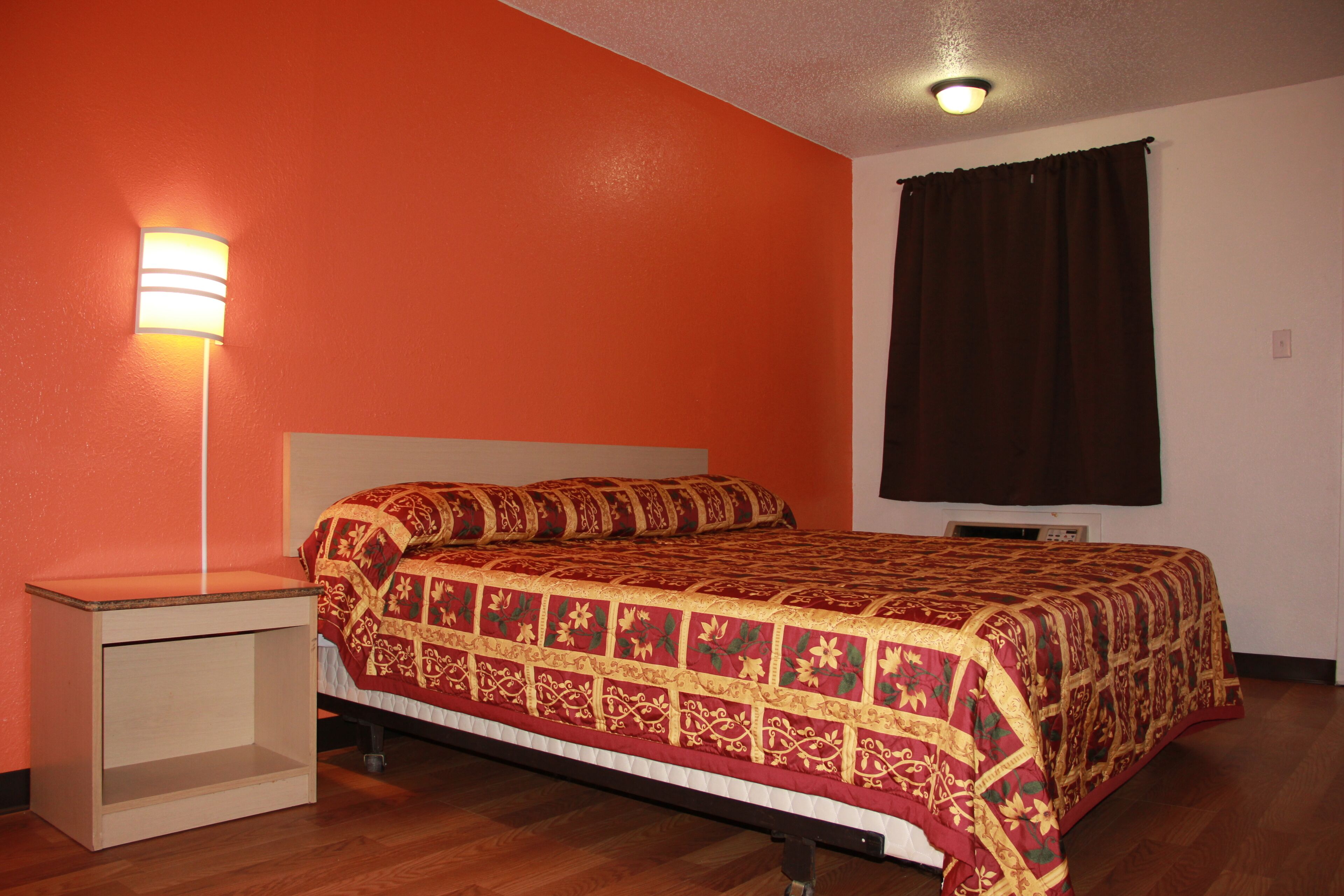 Un dormitorio con cama, mesita de noche, lámpara de pared y ventana con cortinas.