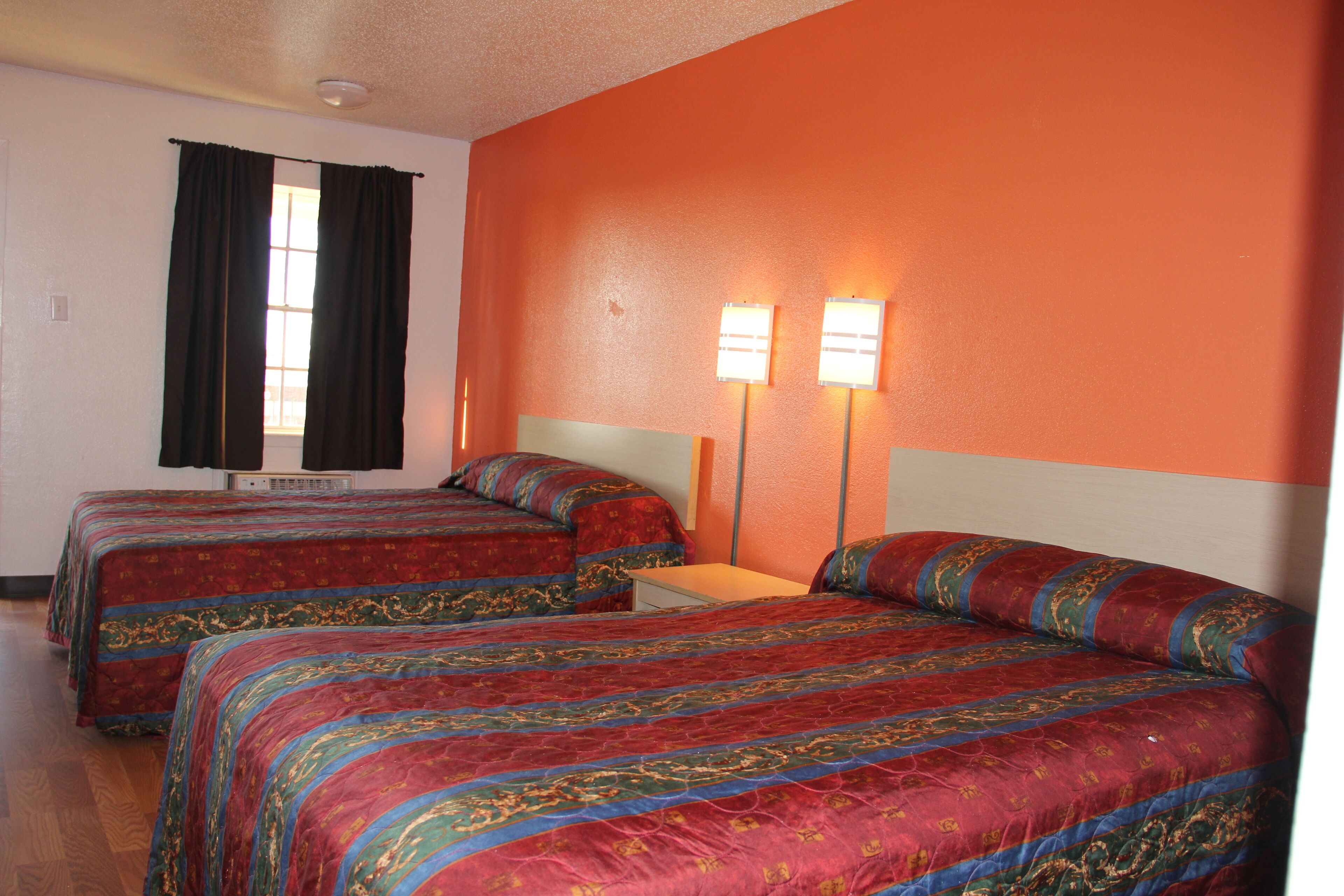 A hotel room with two beds, each with a red and blue patterned bedspread, a window with black curtains, and two wall-mounted lights.