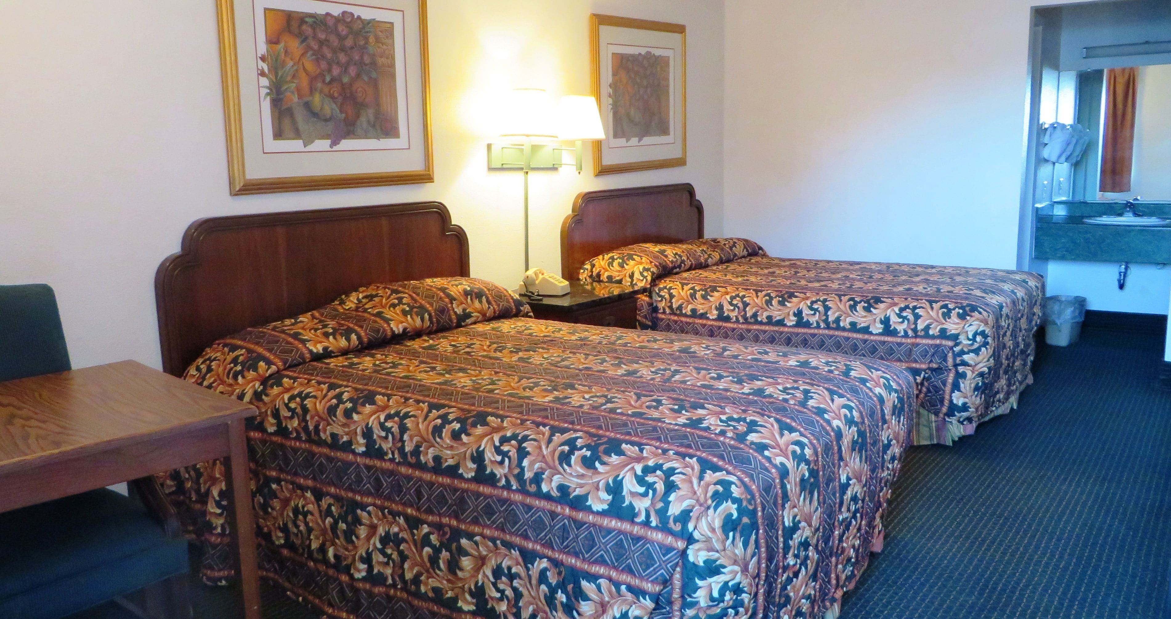 A hotel room with two beds, a desk, a lamp, and a bathroom visible.