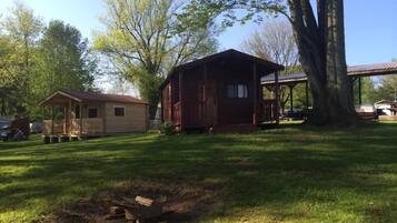 A rustic cabin with a porch and a small shed, surrounded by trees and a grassy yard.
