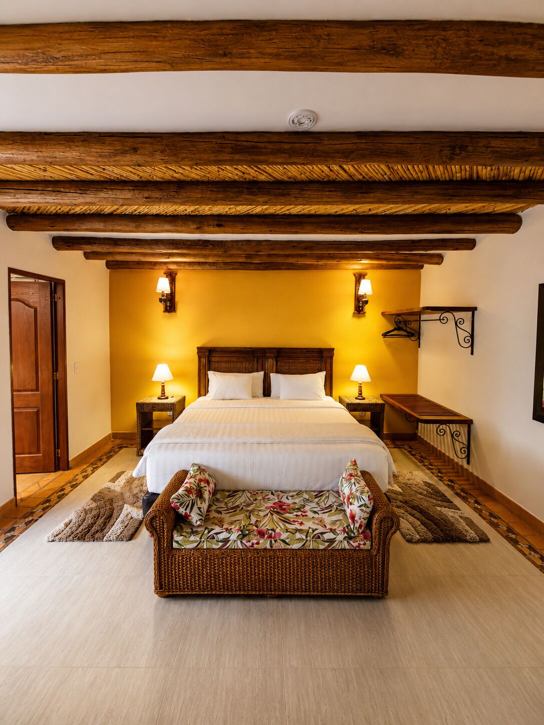 Ein Schlafzimmer mit Holzbalkendecke, einem Bett mit weißer Bettwäsche, zwei Nachttischlampen, einer Bank mit floralen Kissen und einem gemusterten Teppich.