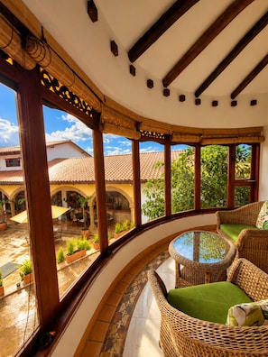 A curved balcony with wicker furniture, a glass table, and a view of a courtyard with plants and a building.