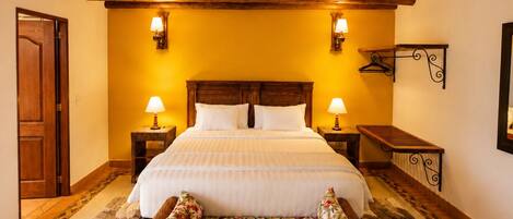 A room with a wooden ceiling, a bed with white linens, two bedside lamps, a wicker sofa, and a wooden bench.