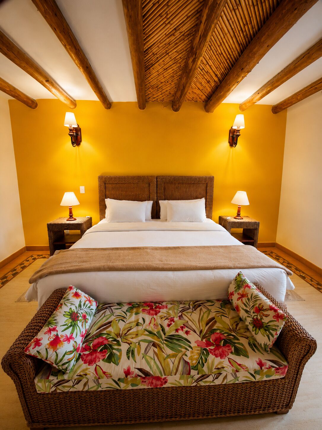 A room with a bed, two nightstands, two wall-mounted lamps, a woven sofa with floral cushions, and a wooden ceiling.