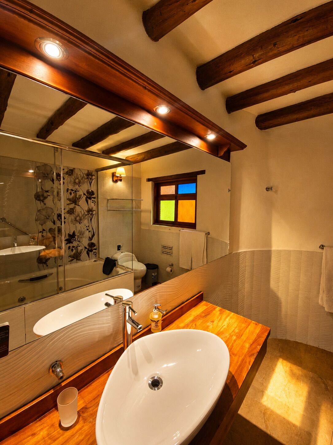 A bathroom with a large mirror, wooden vanity, and a window with colored panes.