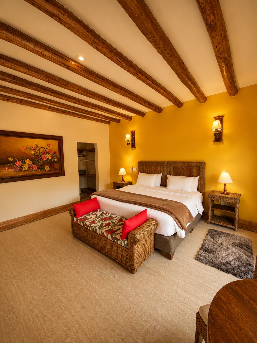 A bedroom with a large bed, two bedside tables with lamps, a wicker sofa with red cushions, a painting on the wall, and wooden beams on the ceiling.