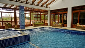 An indoor swimming pool with a tiled floor and walls, surrounded by wicker furniture and large windows.