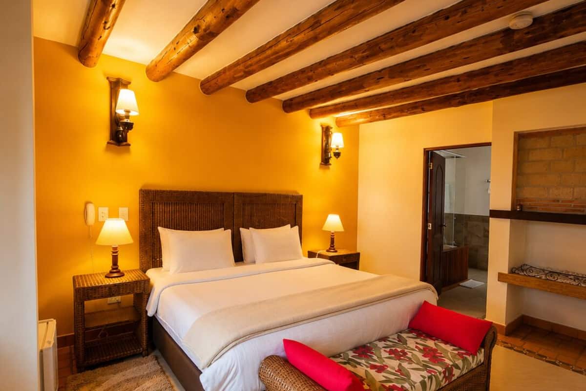 Ein Schlafzimmer mit Holzbalkendecke, einem großen Bett mit weißen Leinen, zwei Nachttischlampen, einer Korbmöbelbank mit rotem Kissen und einer Ziegelwand.