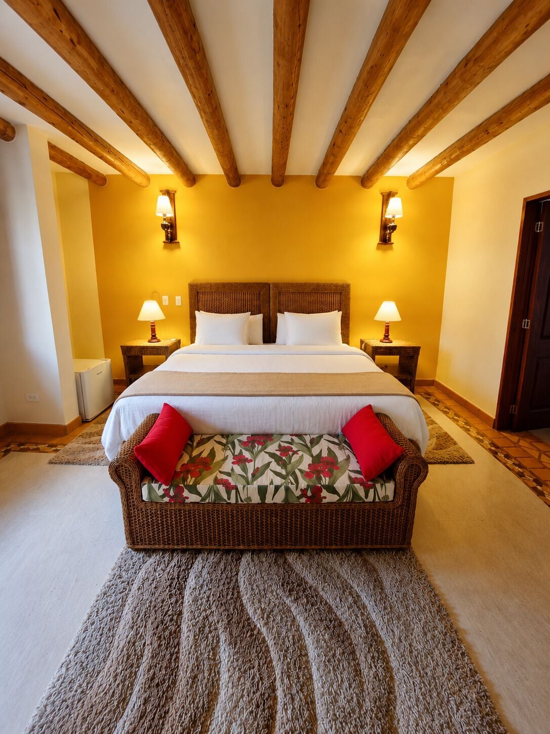 A bedroom with a large bed, wooden beams, two bedside lamps, and a wicker chair with cushions.