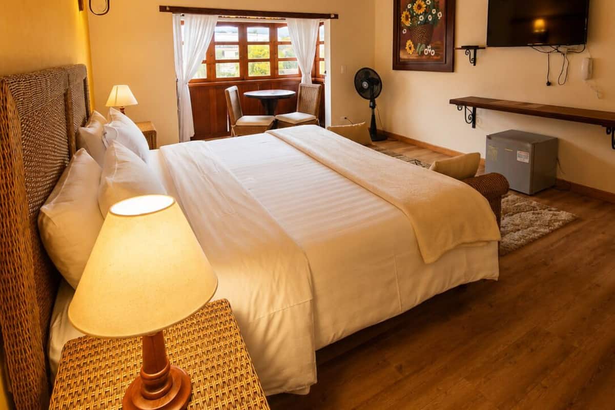 Ein Schlafzimmer mit einem großen Bett, einem Nachttisch mit Lampe, einem an der Wand montierten Fernseher und einem Fenster mit Vorhängen.