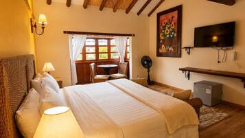Un dormitorio con una cama grande, una mesita de noche con lámpara, un televisor montado en la pared y una ventana con cortinas.