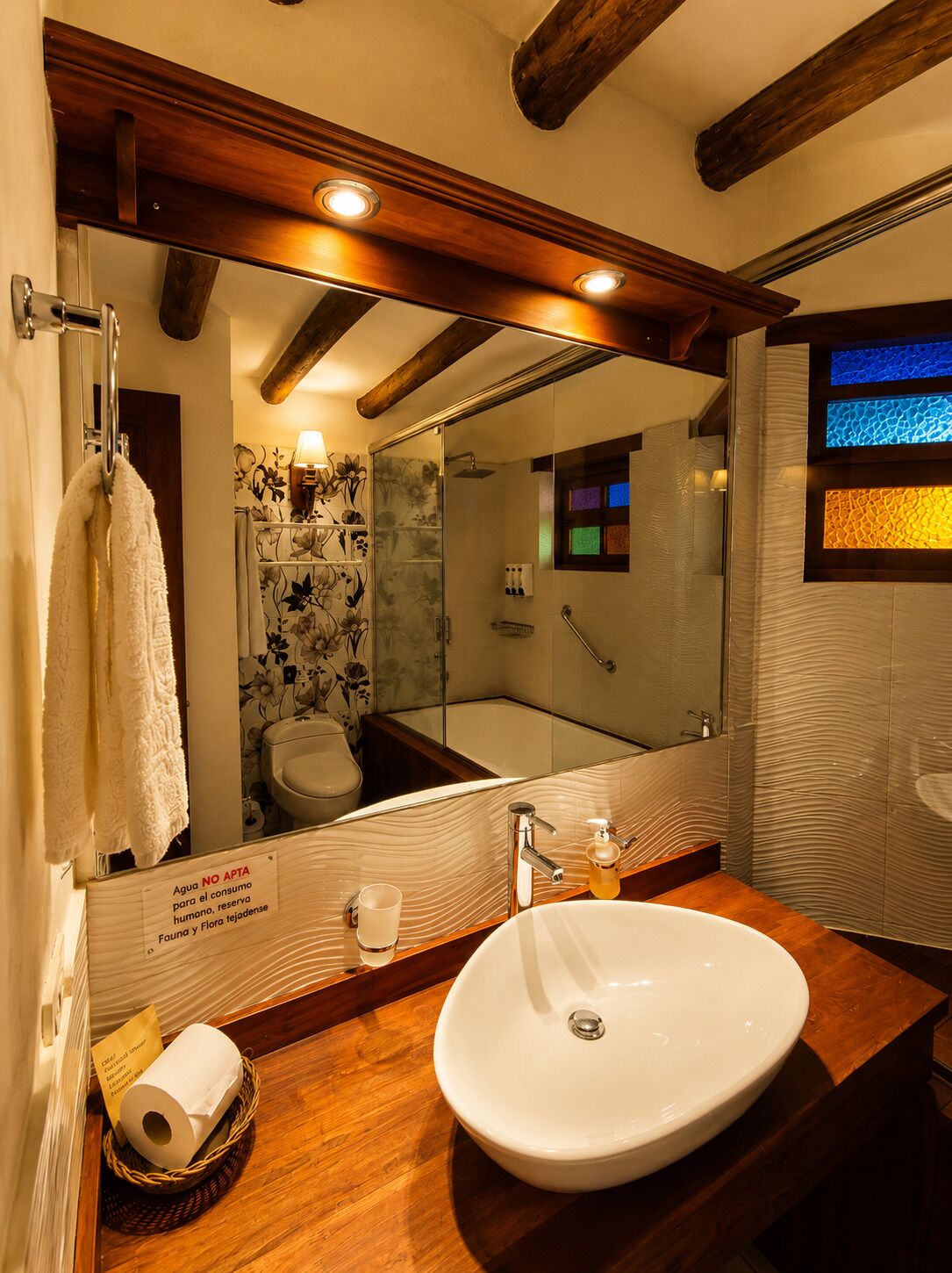 Un baño con un espejo grande, vigas de madera en el techo, un lavabo blanco y una cabina de ducha de vidrio.