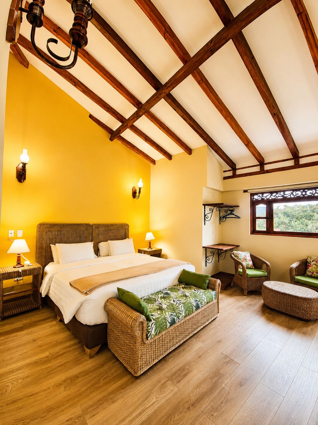 A bedroom with a bed, two nightstands, a bench, wicker furniture, and a ceiling with wooden beams.