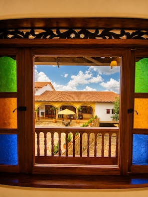 A wooden-framed window with a view of a courtyard featuring a tiled-roof building, outdoor seating, and potted plants.