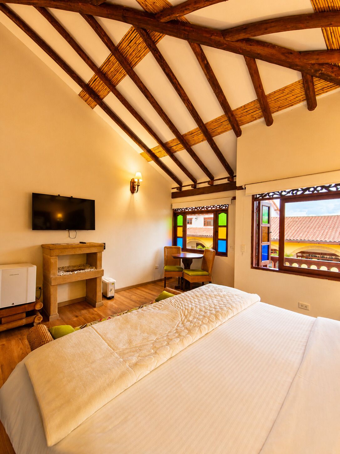 Una habitación con cama, televisión, aparador de madera, una mesita con sillas y ventana con vista a edificios.