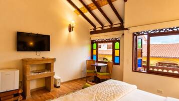 Una habitación con cama, televisión, aparador de madera, una mesita con sillas y ventana con vista a edificios.