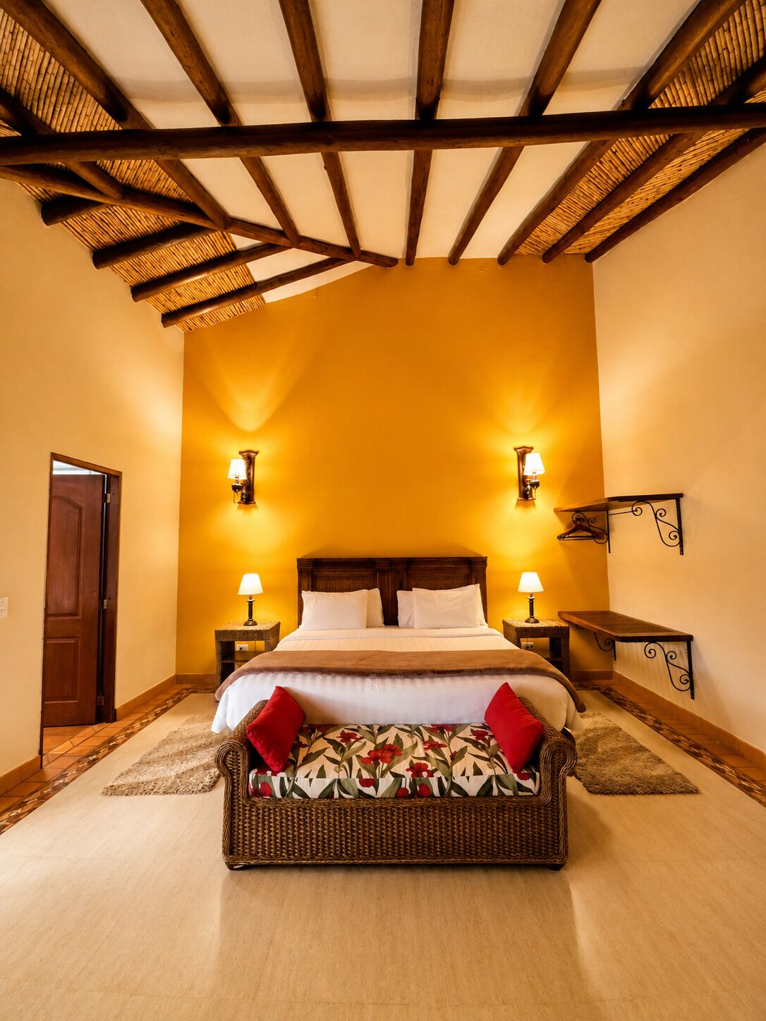 Une chambre d’hôtel avec un plafond en bois, un grand lit avec du linge de lit blanc, deux lampes de chevet, un fauteuil en osier avec des coussins rouges et une petite table avec une lampe.