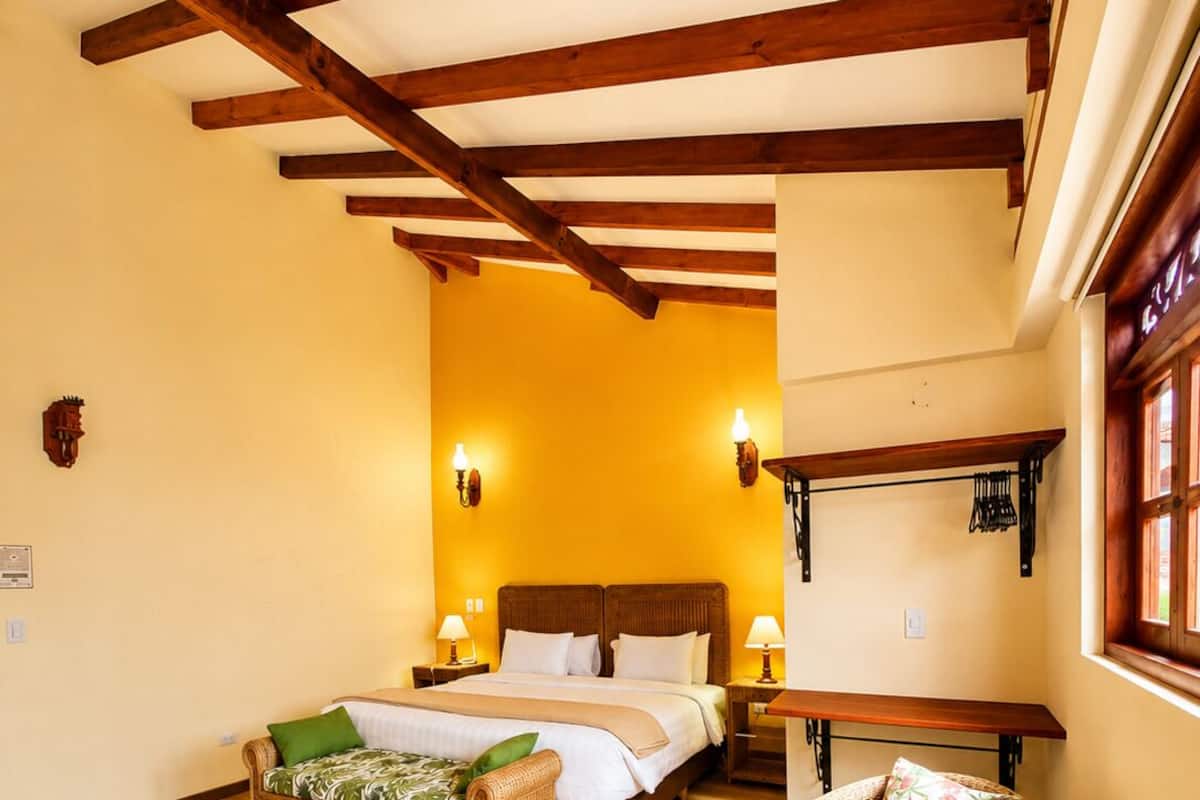 Ein Zimmer mit einem Bett, zwei Nachttischen, einem Korbsessel und einem runden Hocker aus Korb sowie einer Holzbank.