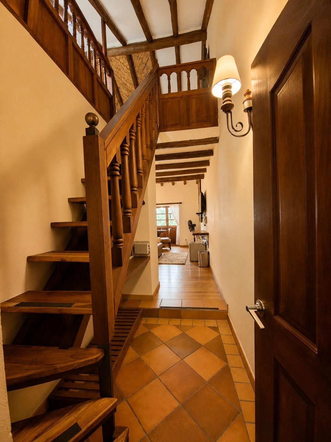 Una escalera de madera con barandilla, una puerta con lámpara y piso de baldosas.