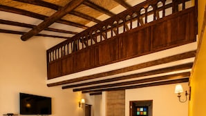 A bedroom with a large bed, a television, a small fridge, a wooden ceiling, and a balcony.