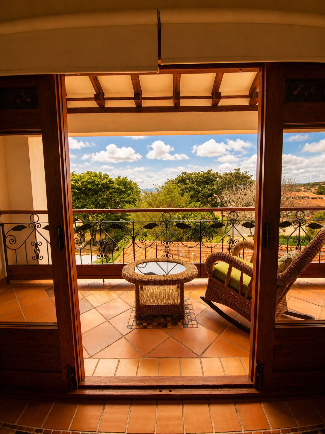 An open balcony with a wicker table and chair, a view of greenery and buildings, and a tiled floor.