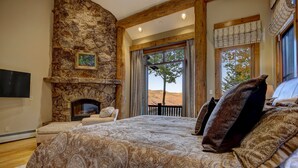 Une chambre avec une cheminée en pierre, un grand lit avec une literie à motifs et une vue sur l’extérieur par une fenêtre.