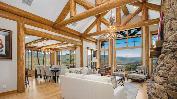 Un vaste espace de vie doté d’un plafond en bois, de grandes fenêtres offrant une vue sur les montagnes, d’une cheminée et d’un coin repas avec une table et des chaises.