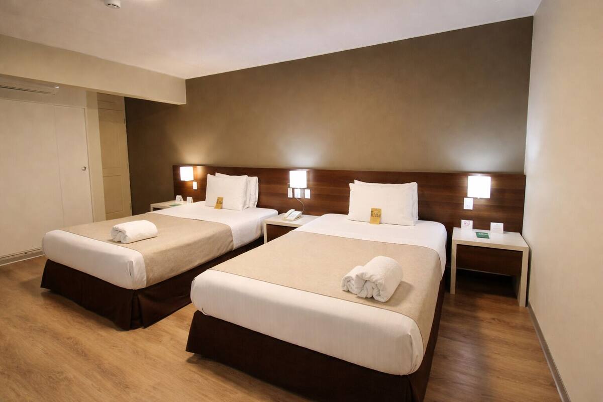 A hotel room with two beds, wooden headboards, bedside tables with lamps, and a door.