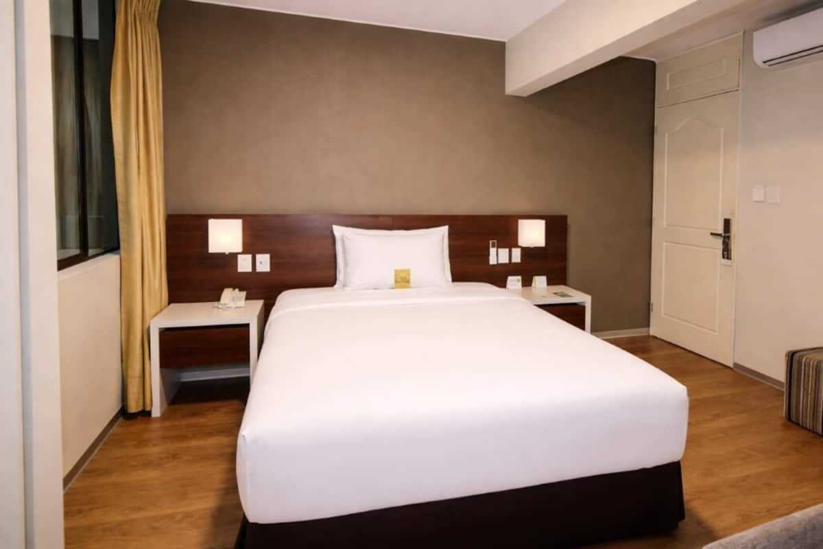 A hotel room with a large bed, bedside tables, a nightstand, a window with curtains, and an air conditioning unit.