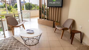 Un vestíbulo moderno de hotel con una gran pantalla de televisión mostrando el nombre del hotel, asientos cómodos, una pequeña mesa y vistas al exterior.