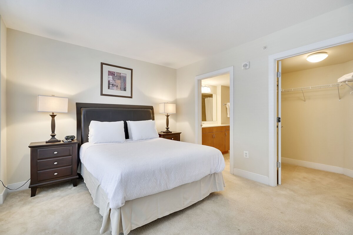 A neatly arranged bedroom with a large bed, two bedside tables with lamps, a framed picture on the wall, and a walk-in closet with a mirror and hanging clothes.