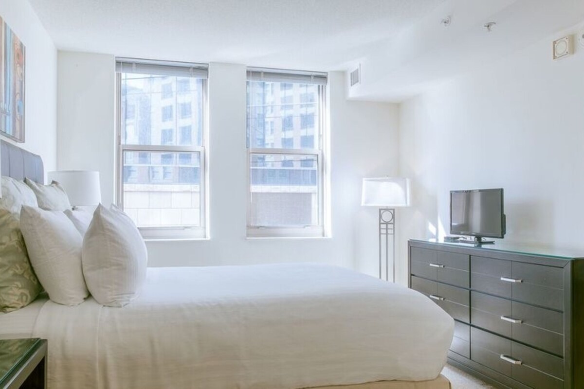 A bedroom with a large bed, two windows, a bedside table with a lamp, a dresser, and a TV.
