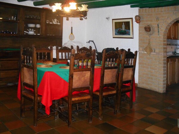 A dining area with a wooden table set for four, surrounded by chairs. A chandelier hangs above the table, and a framed painting is on the wall.