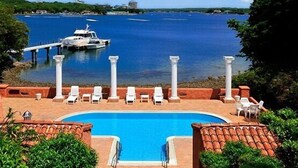 A swimming pool with lounge chairs and a view of a body of water with a dock and boat. - Shima Mediterranean Village (Shima)