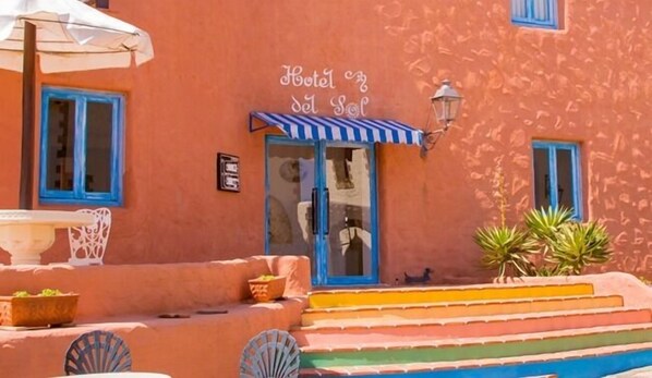 A hotel with a sign reading "Hotel del Sol," featuring a blue awning, a small outdoor seating area with a table and chairs, and a staircase leading to the entrance.