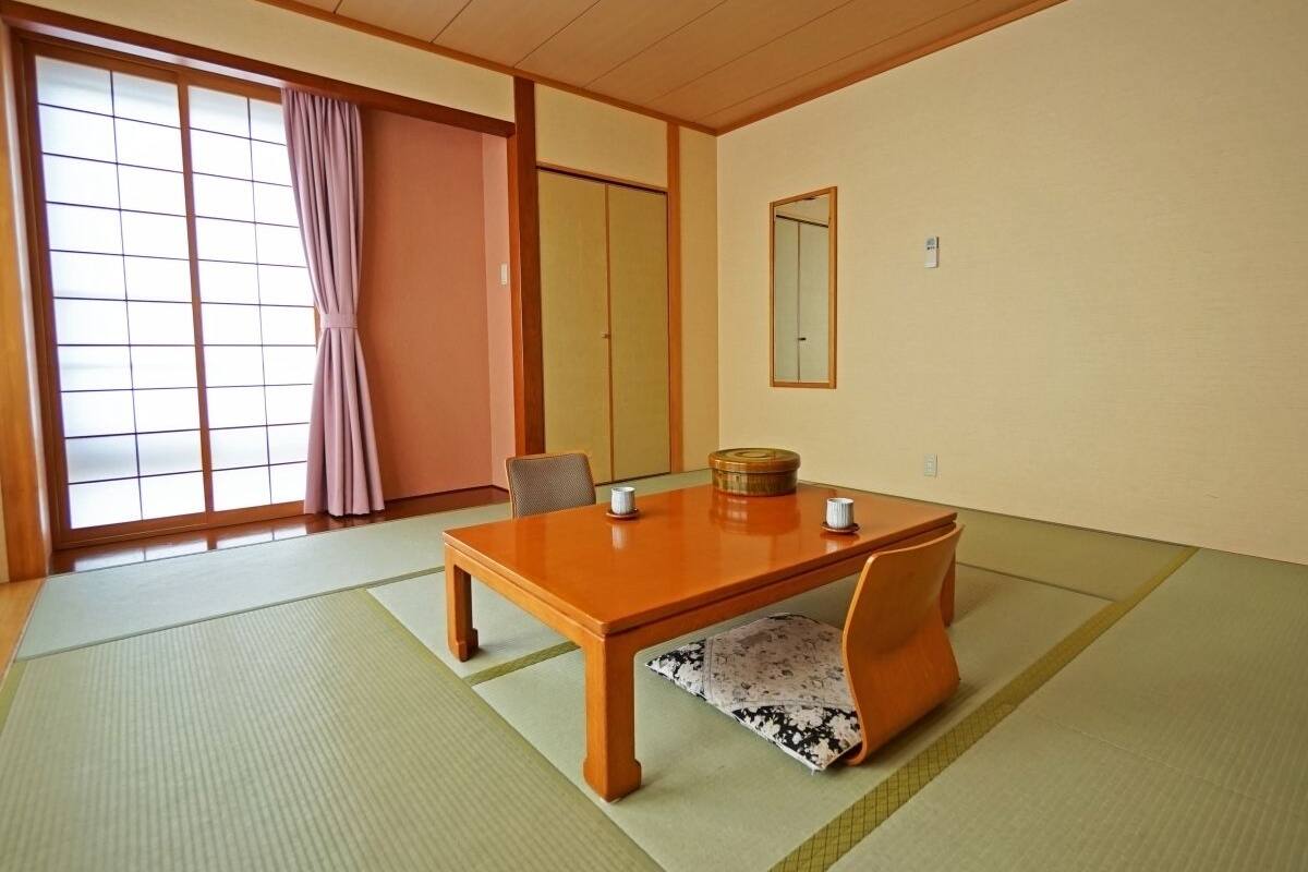 A traditional Japanese room with tatami flooring, a low wooden table, a cushioned bench, sliding doors, and a large window with a view.