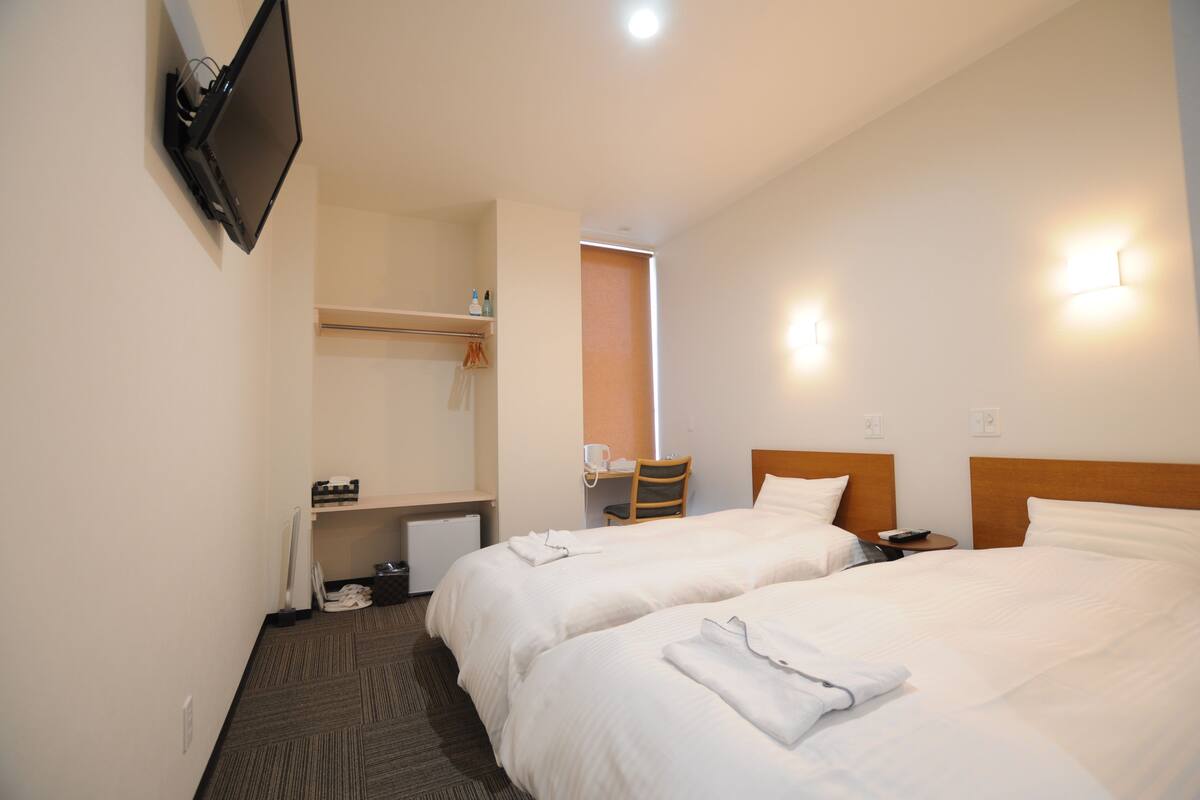 A hotel room with two beds, a TV mounted on the wall, a desk with a chair, and a small table with a phone.