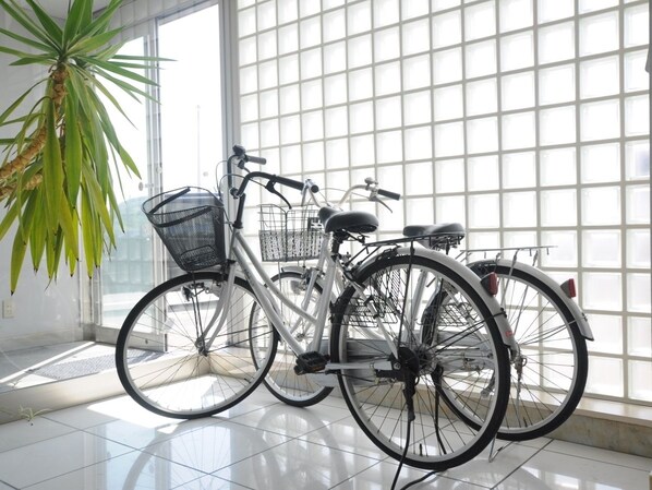 Una bicicleta con canasta y asiento, apoyada contra una pared en una habitación con un ventanal grande y una planta en maceta.