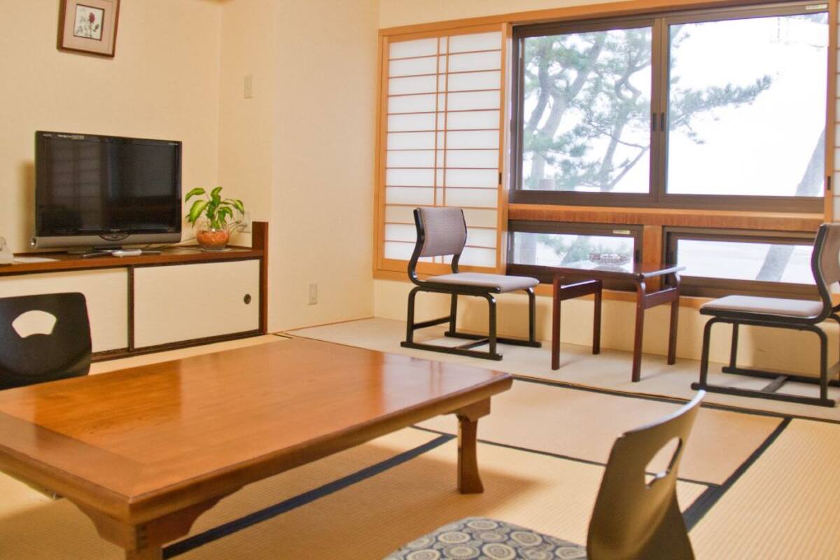 A traditional Japanese-style room with tatami flooring, a wooden table, two chairs, a television, and a window with shoji screens.