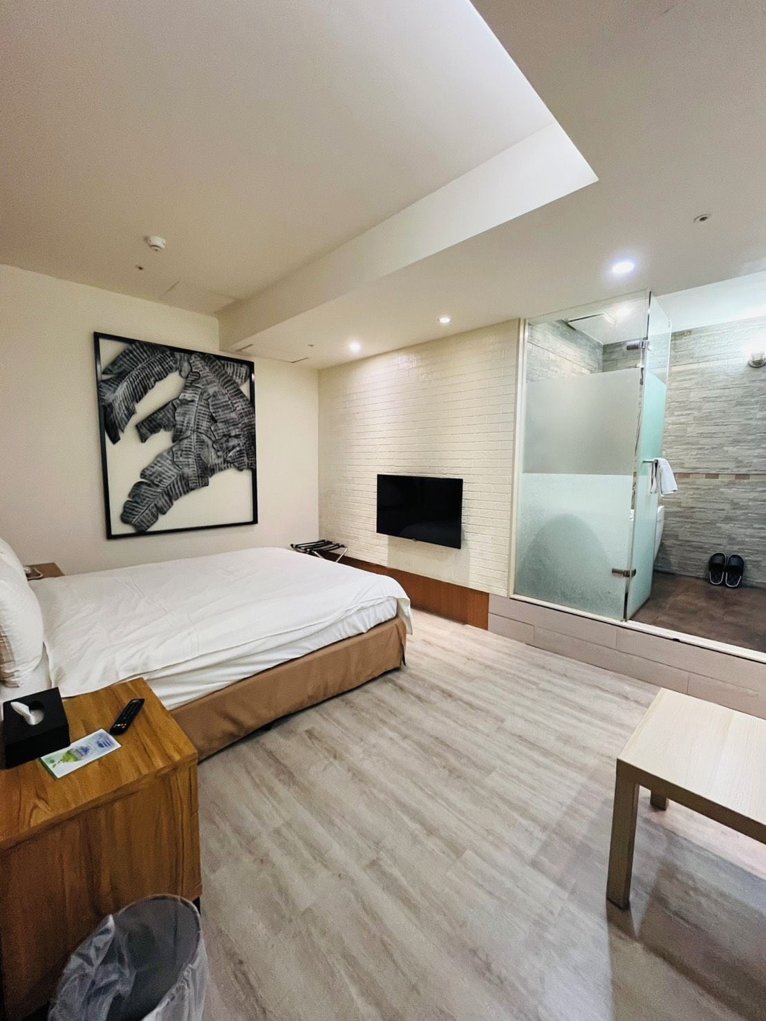 Quarto de hotel moderno com cama, televisão de tela plana na parede, box de vidro para o chuveiro e mesa de cabeceira de madeira com controle remoto.