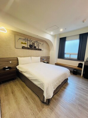 A hotel room with a large bed, a bedside table, a nightstand, a wall-mounted artwork, a window with curtains, and a small desk with a chair.