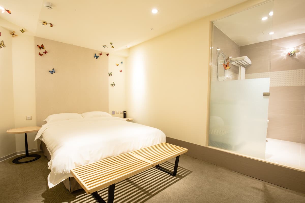 A modern hotel room with a bed, bedside table, bench, and a glass-enclosed shower area.