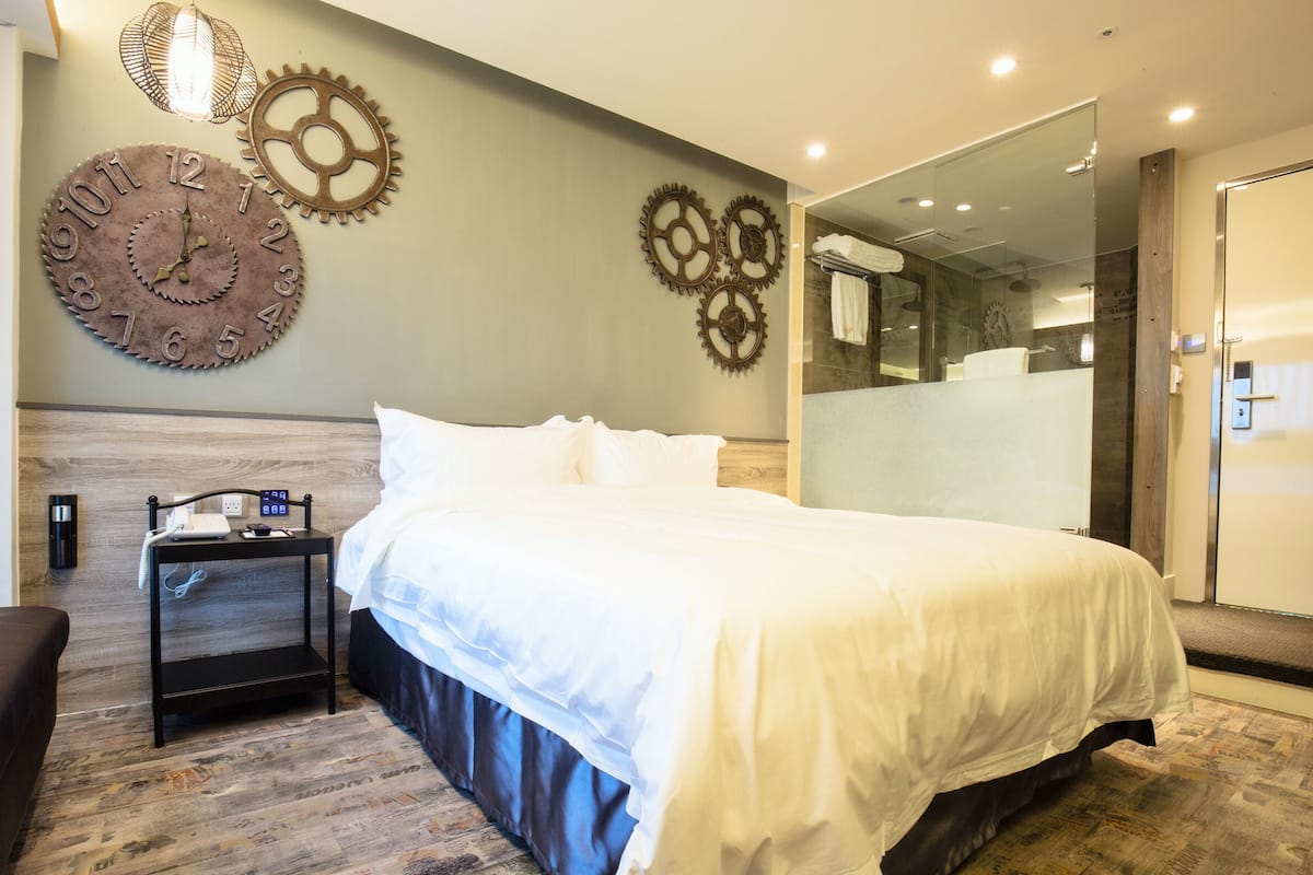 A modern hotel room with a large bed, a nightstand with a phone and a lamp, a wall clock, and a large mirror.